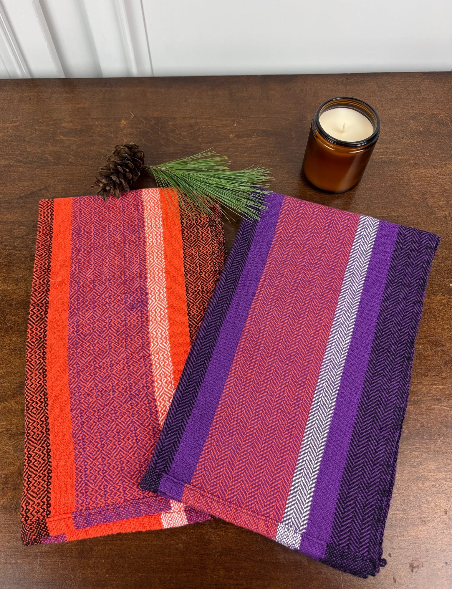 Two woven towels with purple orange black and white stripes on a wooden surface next to a candle.