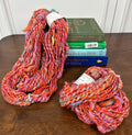 Two skeins of orange and rainbow yarn with a beehive texture on a wooden surface with books stacked behind.