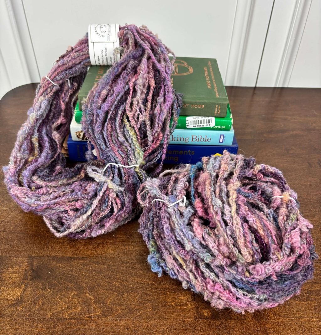 Two skeins of multicolored purple yarn on a wooden surface with books stacked behind.