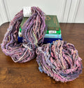 Two skeins of multicolored purple yarn on a wooden surface with books stacked behind.