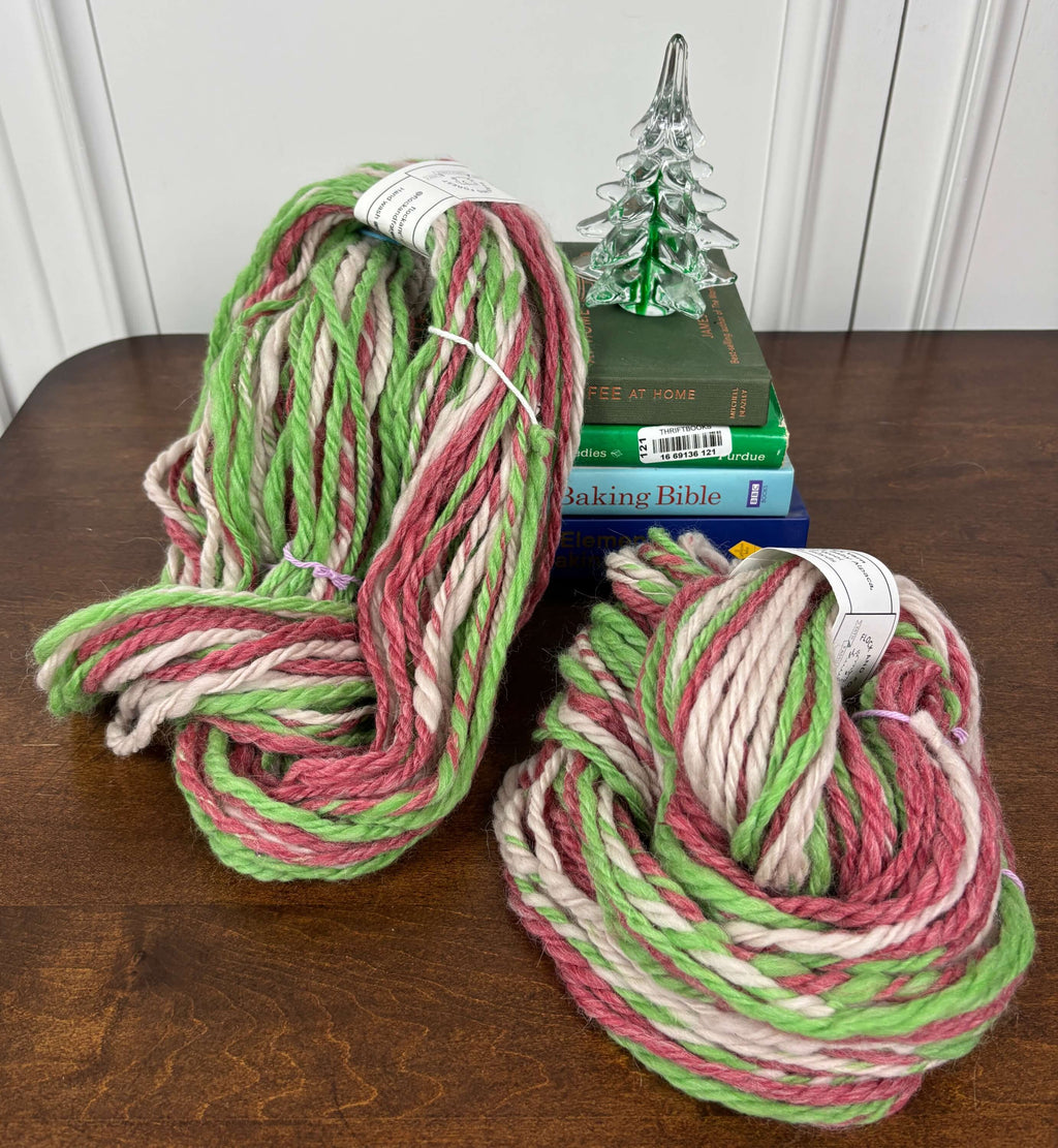 Two skeins of green, red and white yarn on a wooden surface with books and a Christmas tree decoration.