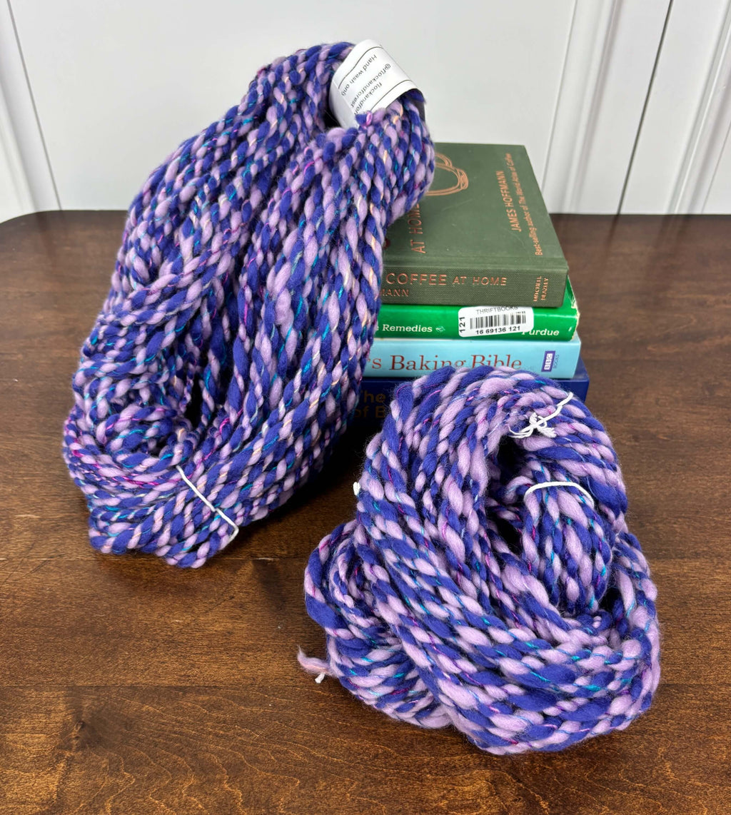 Two dark purple and light purple chunky yarn skeins on a wooden surface with books in the background.